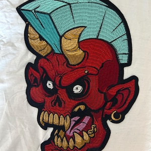 May include: Embroidered patch of a red devil skull with gold horns and teeth, a turquoise mohawk, and a gold earring. The patch has a black border, designed for clothing or accessory application.