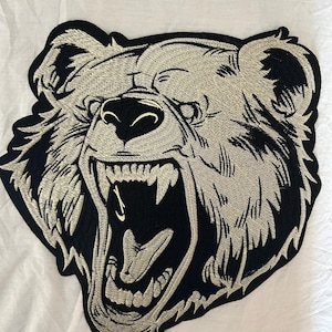 May include: Embroidered patch featuring a roaring bear with a detailed design. The bear's head is outlined in black, with the bear's face and teeth in a light beige color. The patch is likely for jackets, bags, or other apparel.