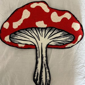 May include: Embroidered patch featuring a red mushroom with white spots. The mushroom has a black outline and a white underside with black lines. The patch is likely for clothing or accessories, adding a whimsical touch.