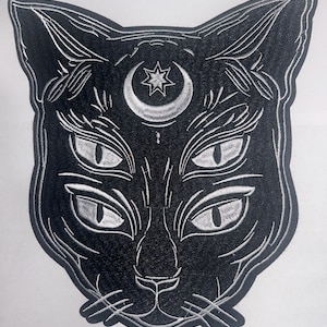 May include: An embroidered patch featuring the head of a black cat with four eyes and a silver crescent moon and star symbol on its forehead. The design is outlined in silver thread against a black background, showing detailed whiskers and ears.