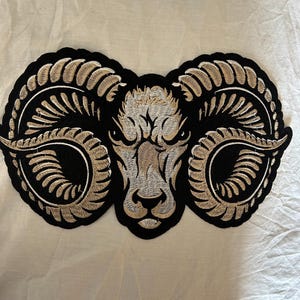Può includere: Toppa ricamata raffigurante una testa di ariete con grandi corna ricurve. La testa dell'ariete è nei toni del beige e del grigio, con un contorno nero. Le corna sono beige con dettagli neri. Per abbigliamento o accessori.