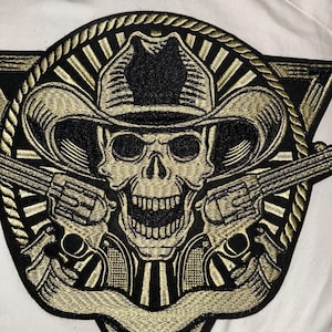 May include: Embroidered patch featuring a skull wearing a cowboy hat, with two revolvers. The design is black and gold, with a triangular border and rope-like edging. The skull has an open mouth.