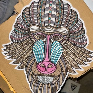 May include: Embroidered patch depicting a detailed baboon face in various colours. The design incorporates brown, pink, blue, and white, with intricate patterns and textures. The patch is suitable for clothing or accessories.