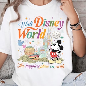 以下が含まれることがあります： ウォルト・ディズニー・ワールドのカラフルなグラフィックが特徴の白いTシャツ。デザインにはミッキーマウス、お城、そして「The happiest place on earth」というフレーズが含まれています。テキストはマルチカラーです。