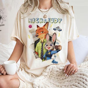 May include: Cream-colored t-shirt featuring the text "NICK & JUDY" above a graphic of Nick Wilde and Judy Hopps from Zootopia. Nick is wearing a green shirt and Judy is in a police uniform. The design includes a police car and newspaper.