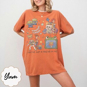 Puede incluir: Una camiseta naranja de manga corta con un estampado gr&aacute;fico de personajes de Toy Story, incluyendo a Woody, Buzz Lightyear y alien&iacute;genas. El dise&ntilde;o incluye el texto Toy Story, Toy Story 5 y You've Got A Friend In Me en un estilo retro colorido.