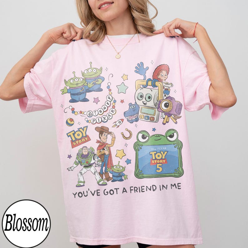 Puede incluir: Una camiseta rosa claro con un estampado gr&aacute;fico colorido de varios personajes de Toy Story, incluidos Woody, Buzz Lightyear y los alien&iacute;genas verdes. El dise&ntilde;o incluye el texto Disney Pixar, Toy Story, Toy Story 5 y You've Got A Friend In Me.