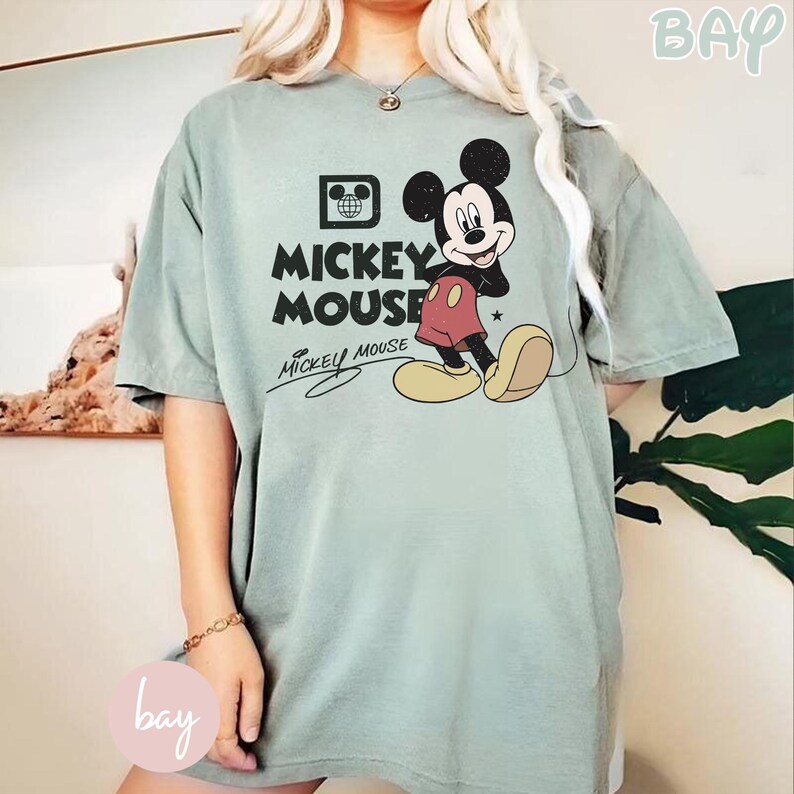 Op de afbeelding: Saliegroen T-shirt met een Mickey Mouse-afbeelding. Het ontwerp bevat de woorden "Mickey Mouse" in het zwart, met Mickey Mouse in zijn klassieke pose. Het shirt heeft een losse pasvorm en is gemaakt van zacht materiaal.