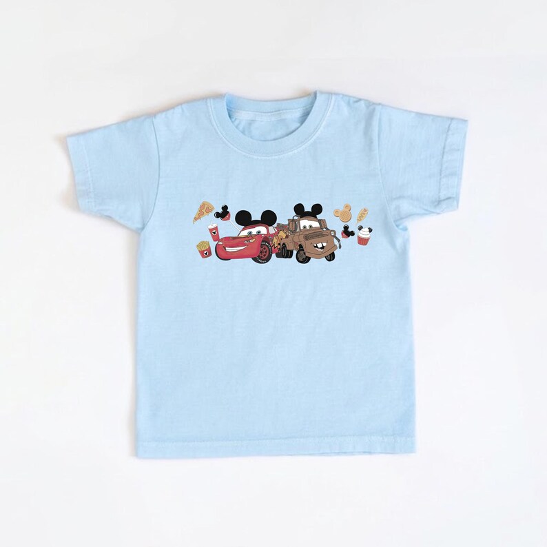 May include: A pale blue children's t-shirt with a cartoon graphic of two cars with Mickey Mouse ears. The red car has a smiling face, and the brown truck has a friendly expression. The design includes cartoon food items.