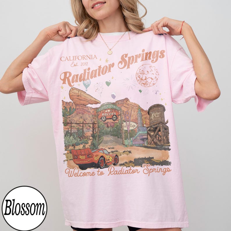 Pu&ograve; includere: T-shirt rosa chiaro con grafica di Radiator Springs, con un'auto da corsa rossa e un paesaggio desertico. Il testo include "CALIFORNIA Est. 2012" e "Welcome to Radiator Springs". La maglietta ha maniche corte e girocollo.