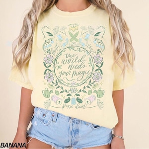 May include: A pale yellow t-shirt featuring a central graphic with the text "the world needs your magic" and "Pixie dust" surrounded by illustrations of flowers, leaves, butterflies, watering cans, spools of thread, and a thimble.