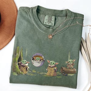 May include: An olive green t-shirt featuring a graphic print of four small green creatures with large ears and tan robes in various poses, including one in a floating pod. The shirt has a crew neck and a Comfort Colors label showing size S-CH.
