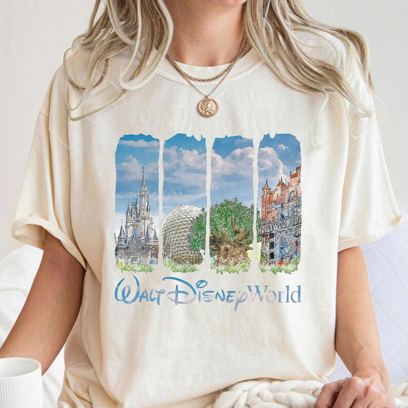 May include: A cream-colored short-sleeve t-shirt featuring a graphic print of four vertical panels depicting iconic theme park landmarks. Below the panels, the text Walt Disney World is printed in a blue script font on the front of the shirt.