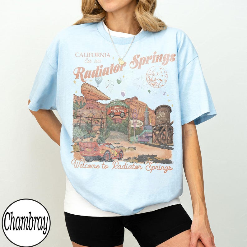Pu&ograve; includere: T-shirt azzurra con grafica di Radiator Springs, con testo che recita "California Est. 2012 Radiator Springs" e "Welcome to Radiator Springs". Il design include un'auto dei cartoni animati e un paesaggio desertico.