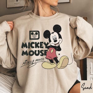 Op de afbeelding: Een beige sweatshirt met een versleten Mickey Mouse-afbeelding. Het ontwerp bevat Mickey Mouses afbeelding, de woorden "Mickey Mouse" en een handtekening. De sweatshirt heeft lange mouwen en een ronde hals.