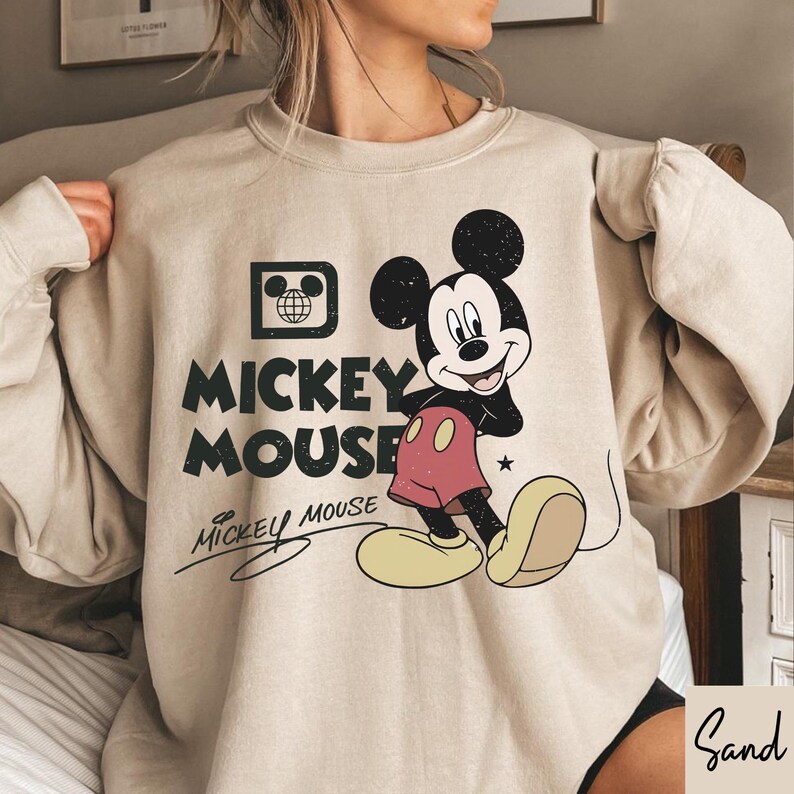 Op de afbeelding: Een beige sweatshirt met een versleten Mickey Mouse-afbeelding. Het ontwerp bevat Mickey Mouses afbeelding, de woorden "Mickey Mouse" en een handtekening. De sweatshirt heeft lange mouwen en een ronde hals.