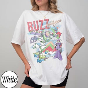 May include: White t-shirt featuring a colorful graphic of Buzz Lightyear, aliens, and Zurg from Toy Story. The design includes the text "BUZZ lightyear" and "LASER BLAST". The shirt has short sleeves and a crew neck.