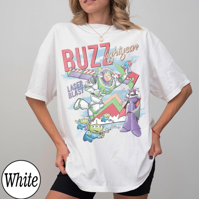 May include: White t-shirt featuring a colorful graphic of Buzz Lightyear, aliens, and Zurg from Toy Story. The design includes the text "BUZZ lightyear" and "LASER BLAST". The shirt has short sleeves and a crew neck.