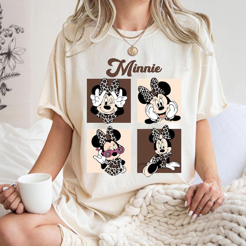 May include: Cream-colored t-shirt featuring a graphic of Minnie Mouse in four different poses. The Minnie Mouse graphic includes a leopard print bow, dress, and accents. The word "Minnie" is printed above the graphic in a brown, cursive font.