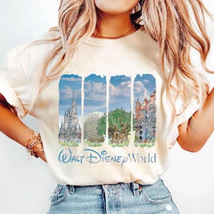 May include: A cream-colored short-sleeve t-shirt featuring a graphic print of four vertical panels depicting Walt Disney World landmarks, including a castle, a geodesic dome, a large tree, and a building, with the text Walt Disney World below.