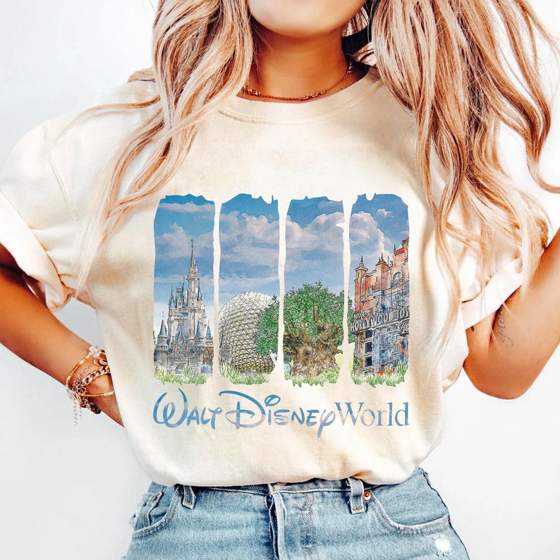 May include: A cream-colored short-sleeve t-shirt featuring a graphic print of four vertical panels depicting Walt Disney World landmarks, including a castle, a geodesic dome, a large tree, and a building, with the text Walt Disney World below.