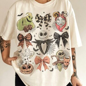 May include: An off-white t-shirt featuring a graphic print of characters and bows from The Nightmare Before Christmas, including Jack Skellington, Sally, Oogie Boogie, Zero, and Lock, Shock, and Barrel, interspersed with spiderwebs and bats.