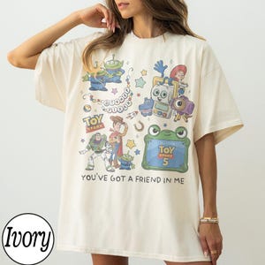 Puede incluir: Una camiseta de manga corta color marfil con un estampado gr&aacute;fico colorido de varios personajes de Toy Story, incluidos Buzz Lightyear, Woody y alien&iacute;genas. El dise&ntilde;o incluye el texto Disney Pixar, Toy Story, Toy Story 5 y You've Got A Friend In Me.