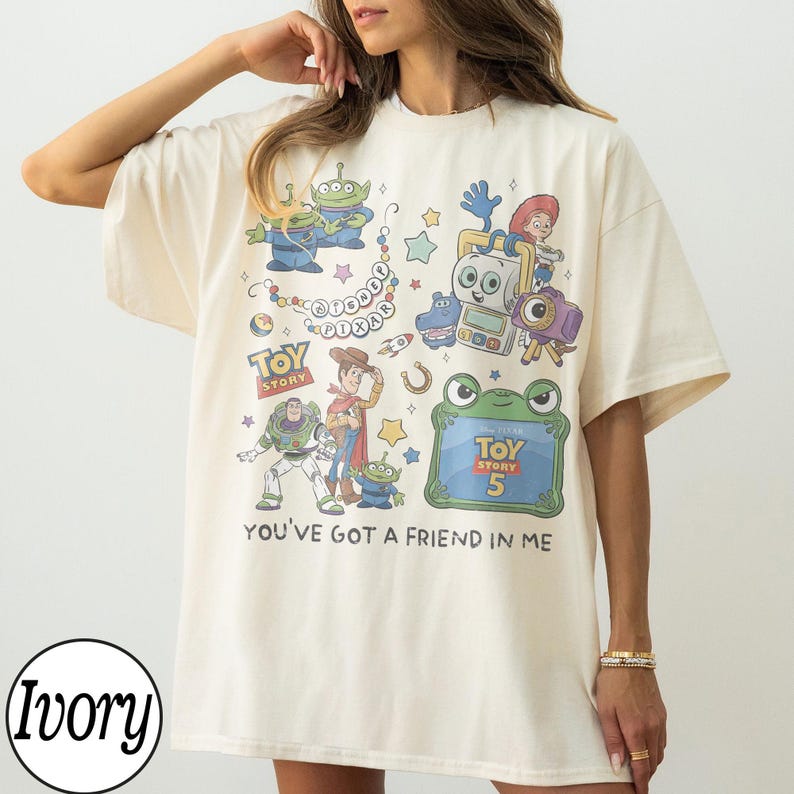 Puede incluir: Una camiseta de manga corta color marfil con un estampado gr&aacute;fico colorido de varios personajes de Toy Story, incluidos Buzz Lightyear, Woody y alien&iacute;genas. El dise&ntilde;o incluye el texto Disney Pixar, Toy Story, Toy Story 5 y You've Got A Friend In Me.