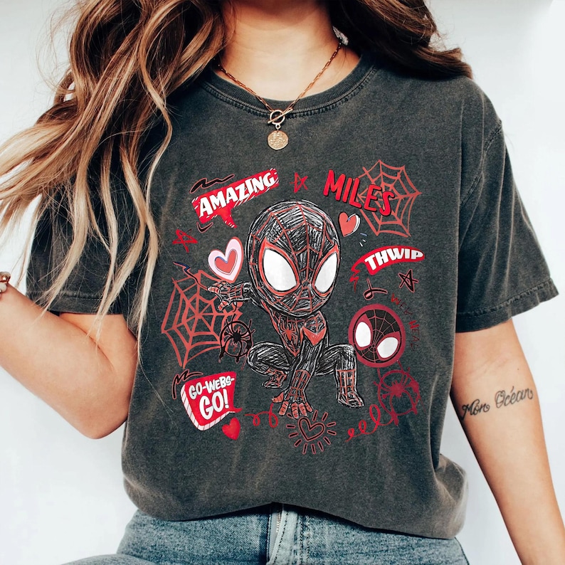 Op de afbeelding: Donkergrijs t-shirt met een cartoon illustratie van een superheld in een zwart en rood pak. Het ontwerp bevat de woorden "Amazing Miles" en "Go-Webs-Go!" in het rood, samen met spinnenwebben en harten.