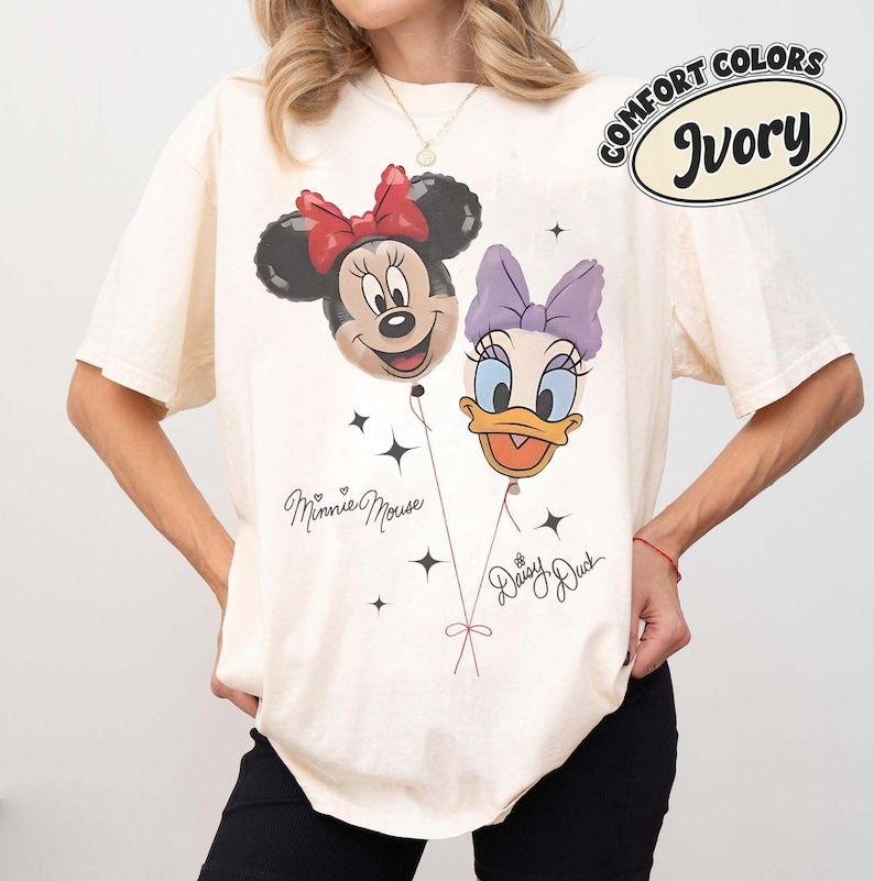 May include: An ivory short-sleeve t-shirt featuring a graphic print of Minnie Mouse and Daisy Duck as balloons. The design includes the text Minnie Mouse and Daisy Duck in cursive script, along with small star accents. A Comfort Colors Ivory label is visible.