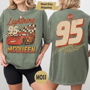 May include: Moss green t-shirt featuring Lightning McQueen from the Cars movie. The front graphic shows McQueen with the number 95 and the text "Lightning McQueen". The back of the shirt has a large "95" and "Lightning McQueen" text.