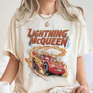 Może przedstawiać: Kremowa koszulka z grafiką Zygzaka McQueena z filmu Auta. Projekt zawiera napis "Lightning McQueen" w kolorze czerwonym i ż&oacute;łtym, z samochodem wyścigowym otoczonym płomieniami.
