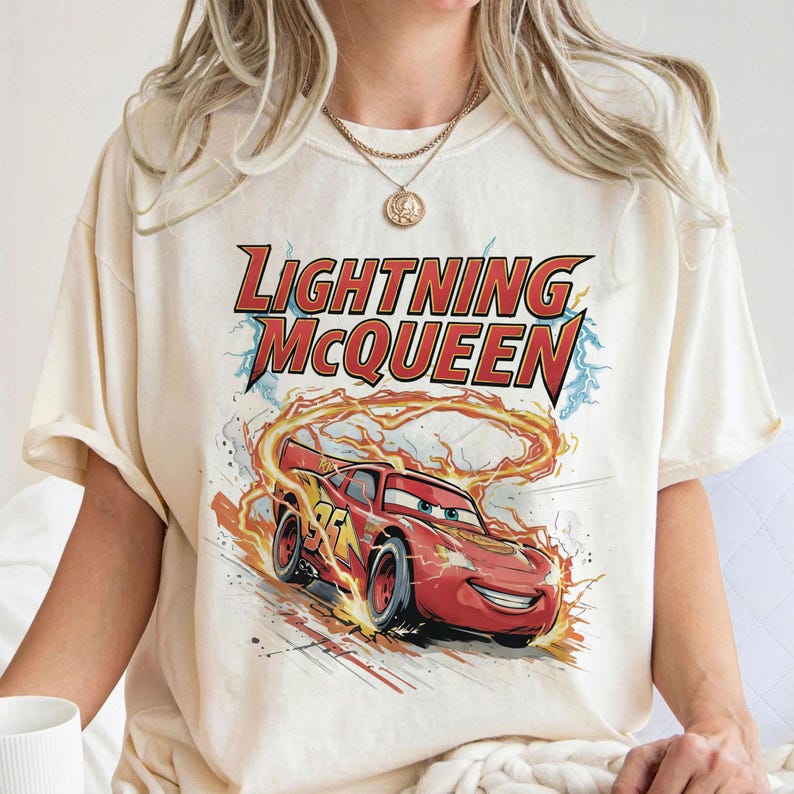 Może przedstawiać: Kremowa koszulka z grafiką Zygzaka McQueena z filmu Auta. Projekt zawiera napis "Lightning McQueen" w kolorze czerwonym i ż&oacute;łtym, z samochodem wyścigowym otoczonym płomieniami.