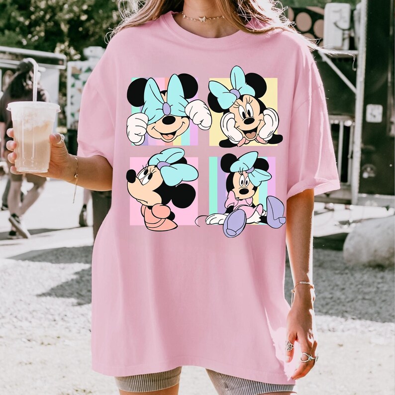 May include: Pale pink t-shirt with a four-panel design of Minnie Mouse. Each panel shows Minnie in different poses, wearing a blue bow. The background of each panel is a different pastel colour, including yellow, pink, and blue.