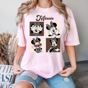 May include: A light pink t-shirt featuring a graphic of Minnie Mouse in four different poses. The word "Minnie" is printed above the graphic in a brown, cursive font. Minnie is wearing a leopard print bow and outfit.