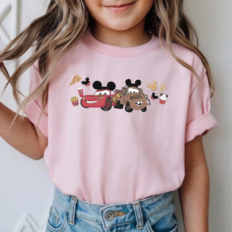 May include: A pale pink t-shirt with a cartoon design of two cars with Mickey Mouse ears, and food illustrations. The cars are red and brown. The shirt is being worn by a child.