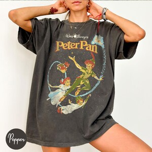 May include: Dark grey t-shirt with a Peter Pan graphic. The design features Peter Pan, Wendy, Tinkerbell, and John, with the text "Walt Disney's Peter Pan" in gold. The shirt has a vintage, worn appearance.