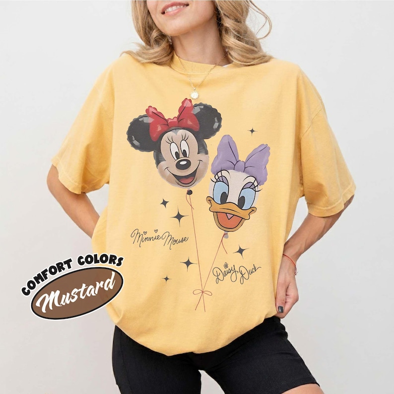 May include: A mustard yellow short-sleeve t-shirt featuring a graphic of Minnie Mouse and Daisy Duck as balloons. The design includes the text "Minnie Mouse" and "Daisy Duck" in cursive script, along with small star accents. The shirt has a crew neck.
