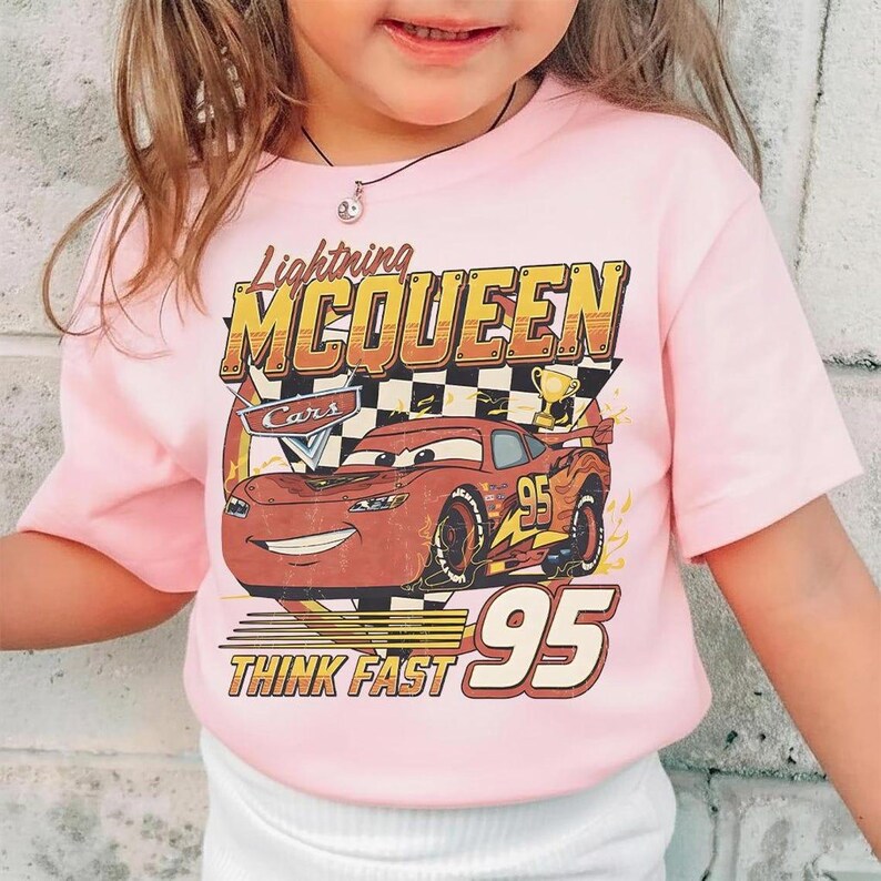 May include: A light pink t-shirt featuring Lightning McQueen from the Cars movie. The graphic includes the text "Lightning McQueen", "Cars", "95", and "Think Fast". The design showcases a red race car with a checkered flag and a trophy.
