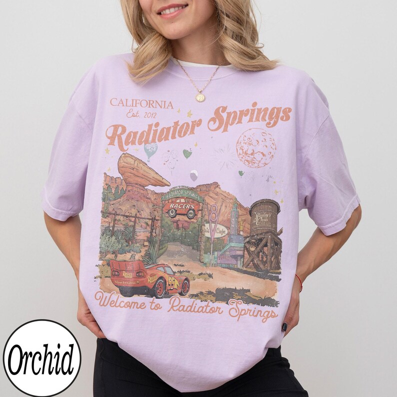 Pu&ograve; includere: Una maglietta lilla chiaro con una grafica di Radiator Springs del film Cars. Il design include un'auto da corsa rossa, uno scenario desertico e il testo "Welcome to Radiator Springs". La maglietta ha anche il testo "California Est. 2017".