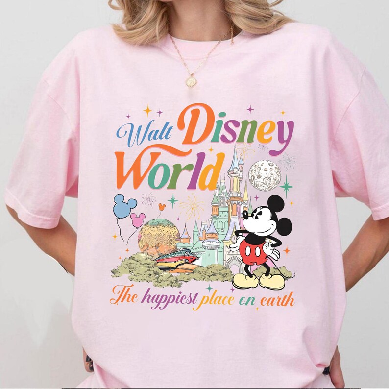 以下が含まれることがあります： ウォルト・ディズニー・ワールドのカラフルなグラフィックが特徴のライトピンクのTシャツ。デザインにはミッキーマウス、お城、風船、そして「The happiest place on earth」というフレーズが含まれています。