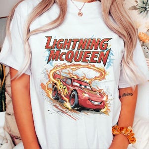 Może przedstawiać: Biała koszulka z grafiką Zygzaka McQueena, czerwonego samochodu wyścigowego z filmu Auta. Tekst "LIGHTNING McQUEEN" znajduje się nad samochodem w kolorze czerwonym i ż&oacute;łtym. Samoch&oacute;d ma numer 95.