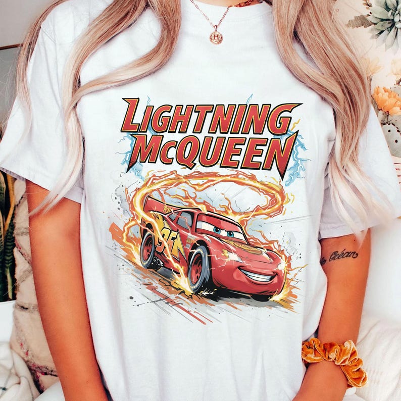Może przedstawiać: Biała koszulka z grafiką Zygzaka McQueena, czerwonego samochodu wyścigowego z filmu Auta. Tekst "LIGHTNING McQUEEN" znajduje się nad samochodem w kolorze czerwonym i ż&oacute;łtym. Samoch&oacute;d ma numer 95.