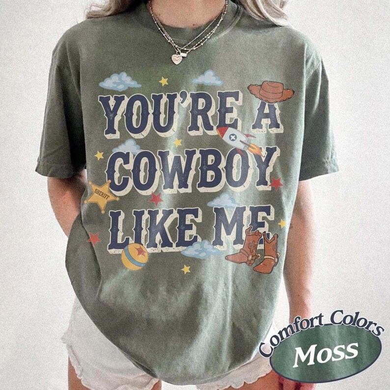 May include: A moss green short-sleeve t-shirt featuring the text "YOU'RE A COWBOY LIKE ME" in dark blue letters. The graphic includes illustrations of a brown cowboy hat, a rocket, a yellow sheriff star, a colorful ball, and small blue clouds.