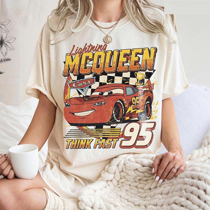 May include: Cream-colored t-shirt featuring a graphic of Lightning McQueen from the movie Cars. The design includes the text "Lightning McQueen", "Cars", "95", and "Think Fast". The shirt has short sleeves and a relaxed fit.