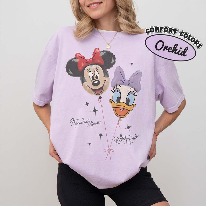 May include: A light purple short-sleeve t-shirt featuring a graphic of Minnie Mouse and Daisy Duck as balloons. The text "Minnie Mouse" and "Daisy Duck" appears below the characters, with a "Comfort Colors Orchid" label in the top right corner.