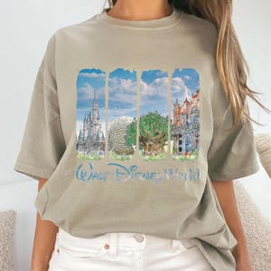 May include: A beige short-sleeve t-shirt featuring a graphic print of four vertical panels depicting Walt Disney World landmarks, including a castle, a geodesic dome, a tree, and a building, with the text Walt Disney World printed below.