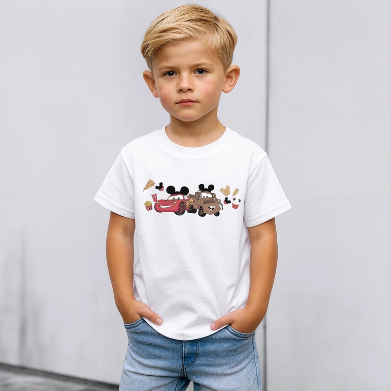 May include: A white t-shirt with a colourful graphic of two cartoon cars with Mickey Mouse ears. The cars are red and brown, with cartoon food icons. The shirt is worn by a young child.