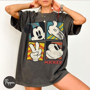 Puede incluir: Camiseta gris oscuro de gran tama&ntilde;o con un gr&aacute;fico desgastado de Mickey Mouse. El dise&ntilde;o incluye cuatro paneles con la cara y la mano de Mickey, y la palabra "Mickey" en rojo. La camiseta tiene un aspecto vintage.