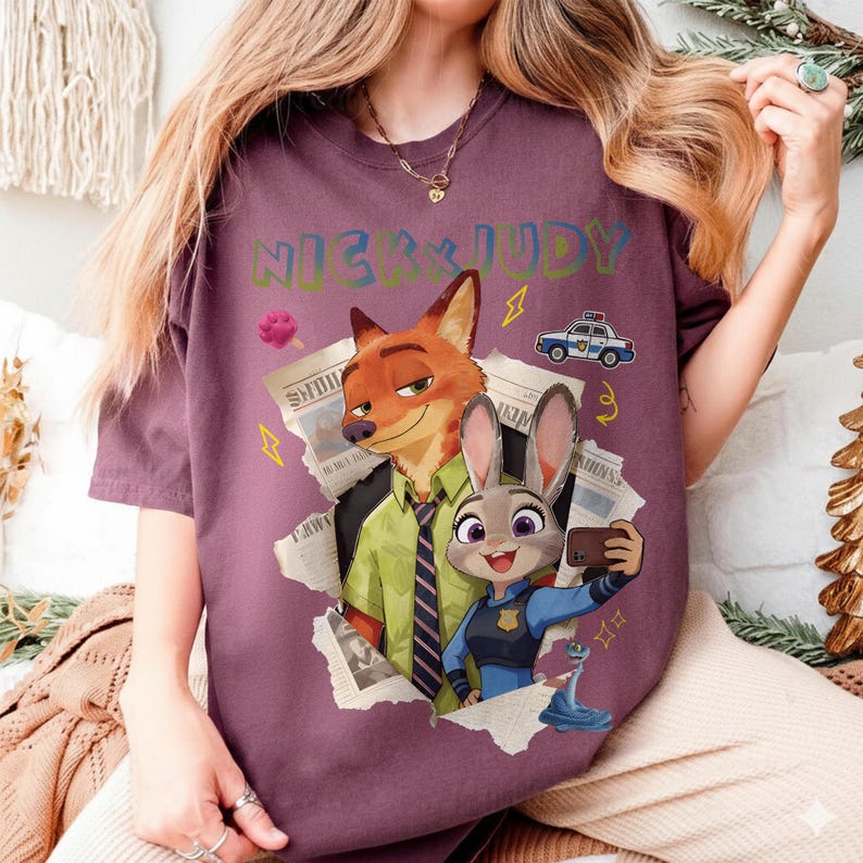 May include: A maroon t-shirt featuring the characters Nick and Judy from Zootopia. The graphic includes the text "NICK JUDY", a police car, newspaper clippings, and a selfie of Judy Hopps. The shirt is a comfortable, casual style.